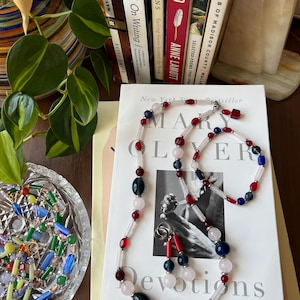 Könnte beinhalten: Eine mehrreihige Halskette mit rosa, roten, blauen und weißen Perlen. Die Halskette liegt auf einem Buch mit dem Titel "Devotions" von Mary Oliver. Das Buch ist ein New York Times Bestseller.