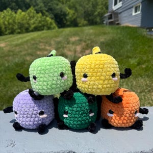 Könnte beinhalten: Fünf kleine, runde, gehäkelte Plüschfiguren in Hellgrün, Gelb, Violett, Dunkelgrün und Orange. Jedes weiche Spielzeug hat eine strukturierte Garnoberfläche, zwei kleine schwarze Perlenaugen, winzige rosa gestickte Wangen und kleine schwarze Gliedmaßen.