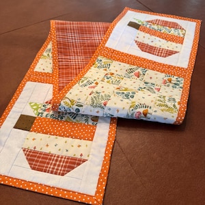 Puede incluir: Un camino de mesa acolchado con bordes de lunares naranjas y blancos, bloques en forma de calabaza con tela de cuadros y flores, y una sección central con fondo crema estampado con pequeños hongos, hojas y motivos botánicos.