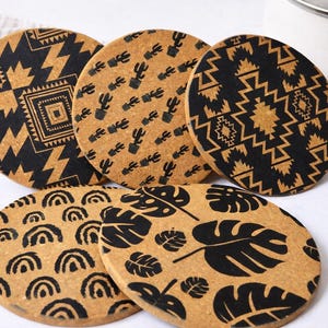 May include: Six round cork coasters with black patterns. Designs include geometric shapes, cacti, arches, and tropical leaves. The coasters are displayed on a light surface, suitable for a home. Each coaster is approximately 10 cm in diameter.