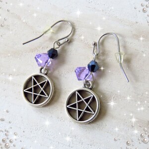 Puede incluir: Pendientes plateados con un colgante de pentagrama. Cada pendiente presenta un grupo de cuentas moradas y azul oscuro sobre el dije. Los pendientes tienen cierre de gancho y protector de oreja transparente.