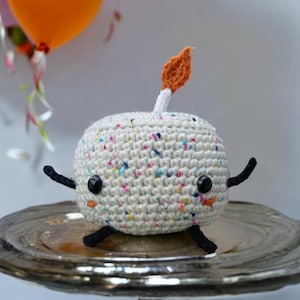 May include: A white crocheted cake with colorful sprinkles and a small orange flame on top. The cake has black eyes and arms and is sitting on a silver cake stand.