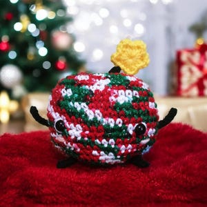 May include: A handmade crochet Christmas ornament in the shape of a round creature with black eyes and arms. The ornament is red, green, and white with a yellow star on top. It is sitting on a red surface, with a Christmas tree in the background.