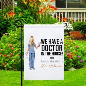 May include: A white garden flag with a cartoon illustration of a female doctor holding a tablet and stethoscope. The text on the flag reads "We have a doctor in the house! Congratulations Dr. Watson".