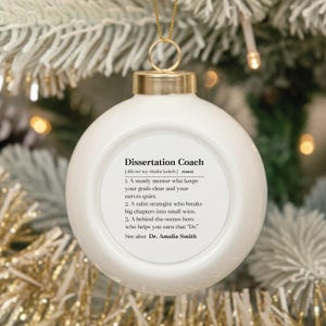 May include: A white, round, ceramic Christmas ornament featuring a gold cap and a gold hanging string. The front displays black text defining the term Dissertation Coach, including three numbered points and the name Dr. Amalia Smith.