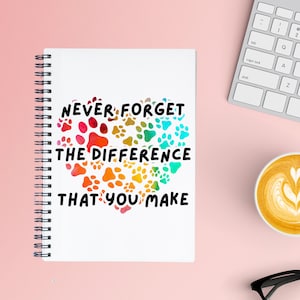 May include: A white spiral bound notebook with a colorful paw print design in the shape of a heart. The text "NEVER FORGET THE DIFFERENCE THAT YOU MAKE" is printed in black on the notebook.