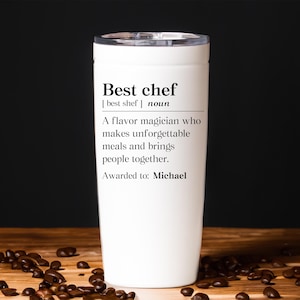 May include: A white insulated tumbler with a black lid. The tumbler has the words "Best chef" printed on it, along with a definition. The tumbler is sitting on a wooden surface with coffee beans scattered around it.