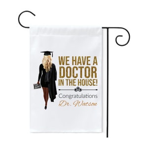 May include: A white garden flag with a black silhouette of a graduate holding a sign that says "I am Done!" The text on the flag reads "We have a Doctor in the house! Congratulations Dr. Watson" in gold glitter lettering.