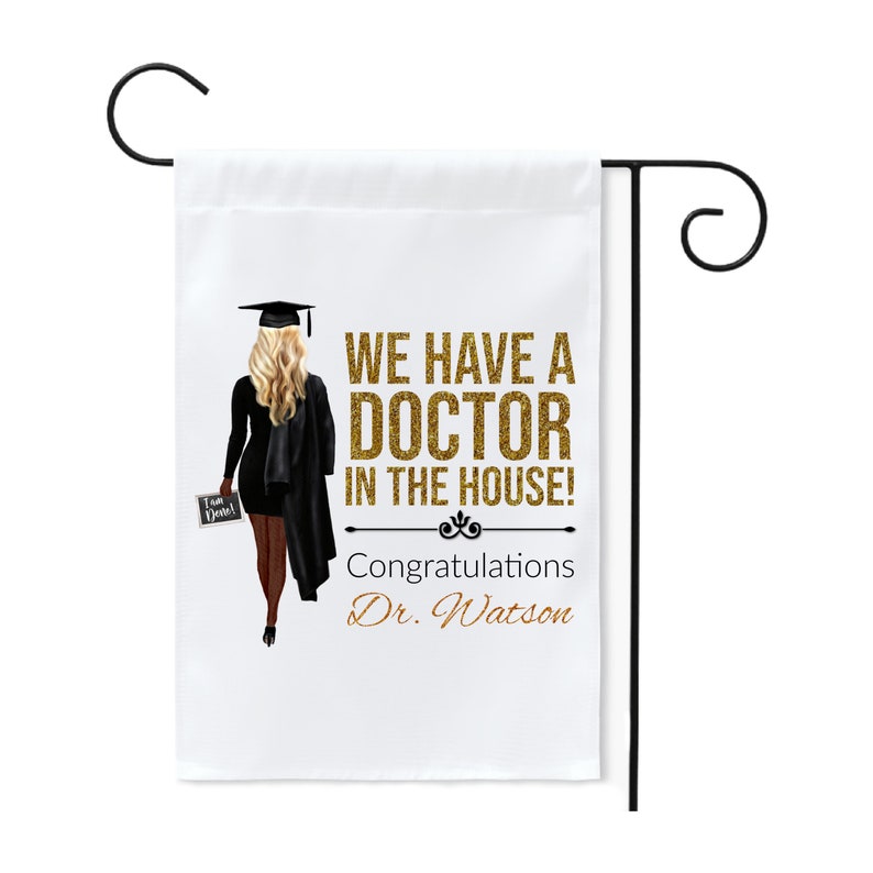 May include: A white garden flag with a black silhouette of a graduate holding a sign that says "I am Done!" The text on the flag reads "We have a Doctor in the house! Congratulations Dr. Watson" in gold glitter lettering.