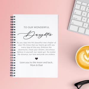 May include: A white spiral-bound notebook with the text "TO OUR WONDERFUL Daughter" and a heartfelt message from Mom & Dad. The notebook is on a pink surface, with a keyboard and a cup of coffee in the background.