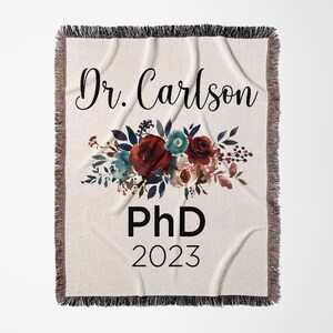 K&ouml;nnte beinhalten: Eine wei&szlig;e Wurfdecke mit einem Blumendesign in Rot-, Rosa- und Blaut&ouml;nen. Der Text "Dr. Carlson PhD 2023" ist in Schwarz auf der Decke aufgedruckt.