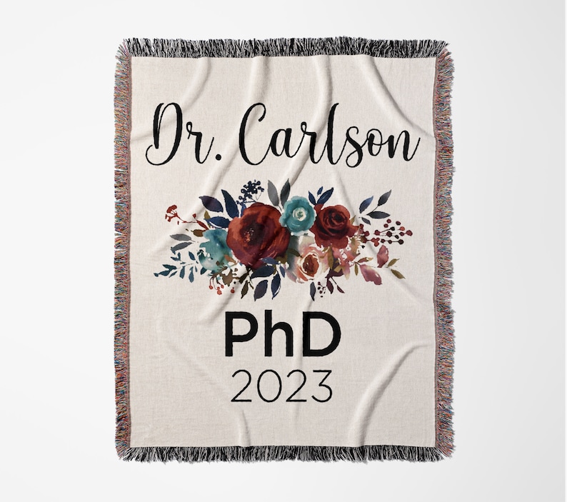 K&ouml;nnte beinhalten: Eine wei&szlig;e Wurfdecke mit einem Blumendesign in Rot-, Rosa- und Blaut&ouml;nen. Der Text "Dr. Carlson PhD 2023" ist in Schwarz auf der Decke aufgedruckt.