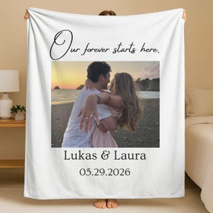 May include: A white fleece blanket featuring a central photograph of a couple embracing on a beach at sunset. The text "Our forever starts here." is printed above the photo, with "Lukas & Laura" and the date "05.29.2026" printed below the image.