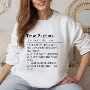 May include: A white long-sleeve sweatshirt featuring black text that defines True Patriots as a noun. The text includes three points: putting country above party, defending every American's vote, and rejecting political violence regardless of the side.