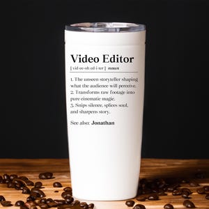 May include: A white travel mug with a black lid, featuring the text "Video Editor" and a definition. The definition describes the role of a video editor. The mug is on a wooden surface with coffee beans.