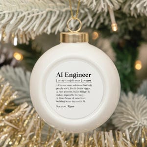 May include: A round white ceramic Christmas ornament featuring a gold metal cap and a gold hanging string. The front displays black text defining AI Engineer as a noun, including three numbered points and the phrase See also: Ryan.