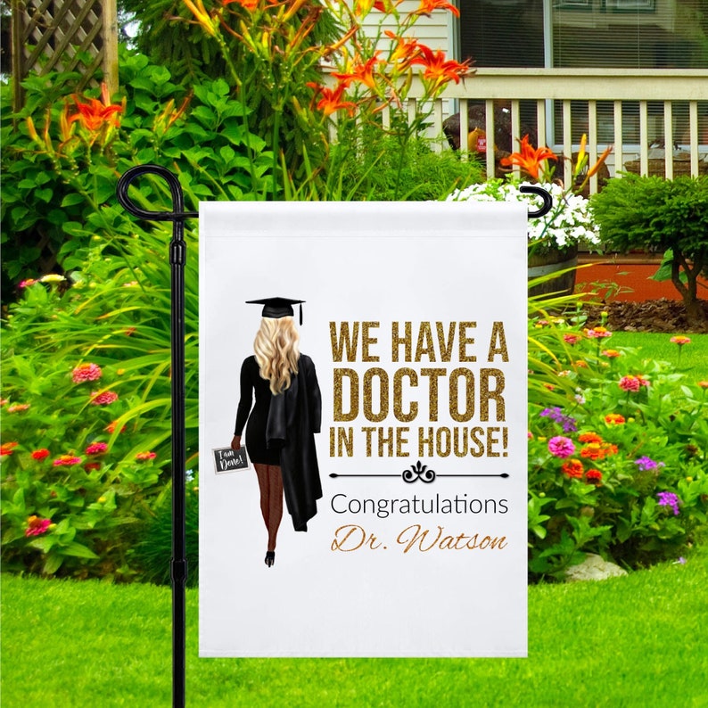 May include: A white garden flag with a gold glitter design. It features a woman in a black graduation gown and cap, holding a diploma. The text reads "We have a doctor in the house! Congratulations Dr. Watson".