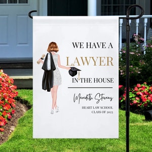 May include: A white lawn sign with a black and white illustration of a woman in a graduation gown and cap. The text on the sign reads "We have a lawyer in the house" with the name "Meredith Stevens" and "Heart Law School Class of 2023" below.