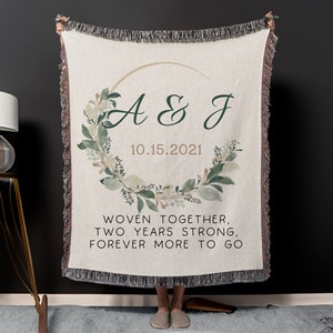 May include: A woven throw blanket with a green and white floral wreath design. The wreath encircles a gold circle with the initials "A & J" and the date "10.15.2021". The text "WOVEN TOGETHER, TWO YEARS STRONG, FOREVER MORE TO GO" is printed below the wreath.