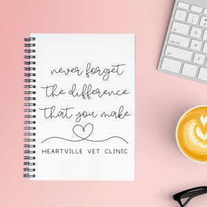 May include: A white notebook with a black spiral binding. The notebook has a quote that reads "never forget the difference that you make" with a heart symbol and the text "HEARTVILLE VET CLINIC".