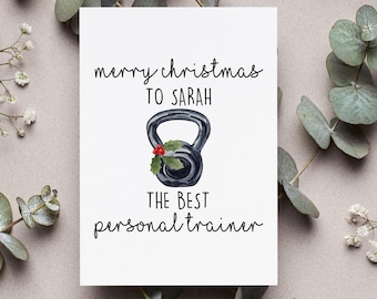 May include: A white greeting card with the text "merry christmas TO SARAH" and "THE BEST personal trainer". The card features a watercolor illustration of a black kettlebell decorated with holly. Eucalyptus and small white flowers surround the card.