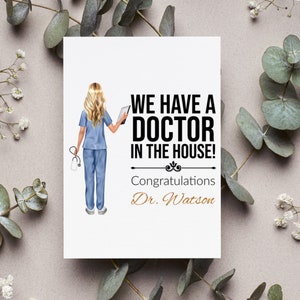 May include: A white card with a black and white illustration of a female doctor in scrubs holding a clipboard. The text "We have a doctor in the house!" is in black and the text "Congratulations Dr. Watson" is in gold.