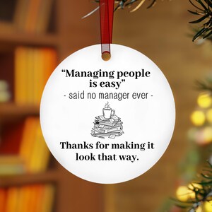 May include: A round white ceramic ornament hanging from a red ribbon. The text reads: "Managing people is easy" - said no manager ever - followed by a line drawing of a coffee mug on a stack of papers, and "Thanks for making it look that way."