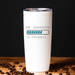 May include: White travel mug with a black and teal design. The text on the mug reads "Dr. Johnson In Progress..." with a progress bar graphic.