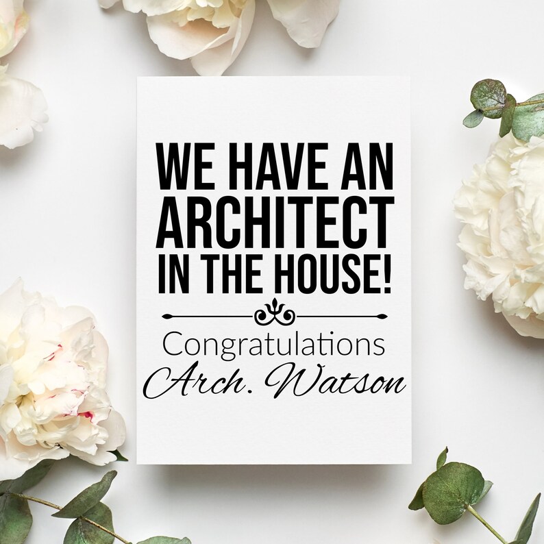 May include: A white card with black text that reads "We have an architect in the house!" with a decorative flourish and the text "Congratulations Arch. Watson". The card is surrounded by white flowers and green leaves.