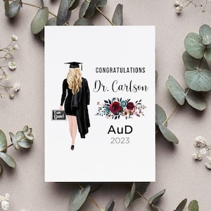 May include: Congratulations Dr. Carlson, Aud 2023. A graduation card with a black and white illustration of a woman in a cap and gown holding a chalkboard that says "I am Done!"  The card has a floral design with red, pink, and blue flowers.