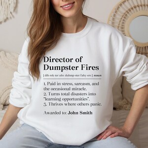 May include: White sweatshirt with the text "Director of Dumpster Fires" in black. The definition includes phrases like "Paid in stress, sarcasm, and the occasional miracle." and "Thrives where others panic." The name "John Smith" is listed below.
