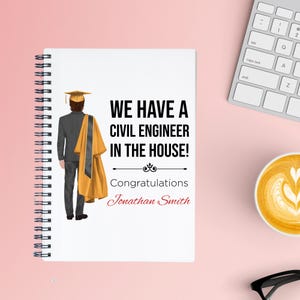May include: A spiral-bound notebook with the text "WE HAVE A CIVIL ENGINEER IN THE HOUSE!" and "Congratulations Jonathan Smith." An illustration of a graduate in a cap and gown is on the left. The background is a light pink.