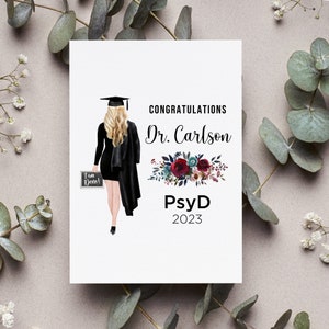 May include: Congratulations Dr. Carlson PsyD 2023 graduation card with a black and white illustration of a woman in a cap and gown holding a chalkboard that says "I am Done!" and a floral design with red, pink, and blue flowers.