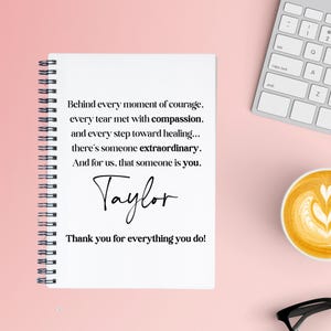 May include: A white spiral-bound notebook with black text that reads, "Behind every moment of courage...Taylor...Thank you for everything you do!" A keyboard, a cup of coffee, and glasses are also visible.