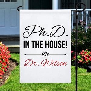 May include: White garden flag with black text that reads "Ph.D. in the house!" and "Dr. Wilson" in red text.