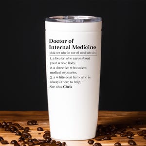 May include: A white insulated tumbler with a silver lid. The tumbler features the text "Doctor of Internal Medicine" with a definition. The tumbler is on a wooden surface with coffee beans.