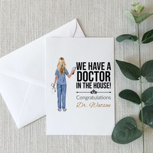 May include: A white card with a black and white illustration of a woman in a blue medical uniform holding a tablet. The text on the card reads "We have a Doctor in the house! Congratulations Dr. Watson."