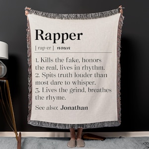 May include: Cream-colored throw blanket with black fringe, featuring the definition of "Rapper" in large black text. The definition includes three points and the phrase "See also: Jonathan."