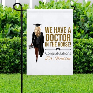 May include: A white garden flag with a black silhouette of a person wearing a graduation cap and gown. The text on the flag reads "We have a Doctor in the house! Congratulations Dr. Watson".