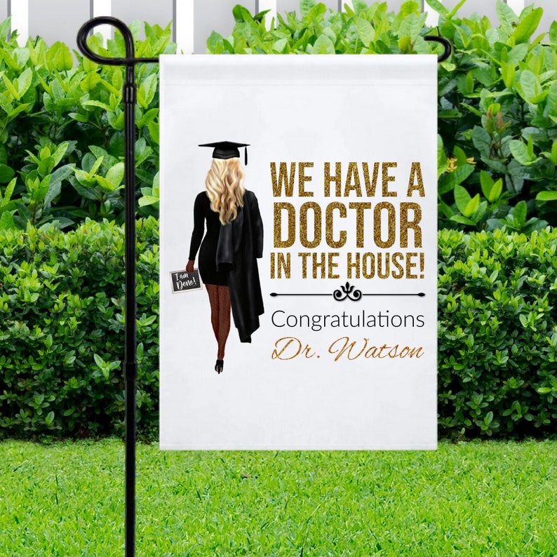 May include: A white garden flag with a black silhouette of a person wearing a graduation cap and gown. The text on the flag reads "We have a Doctor in the house! Congratulations Dr. Watson".
