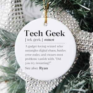 May include: A white, round Christmas ornament with the text "Tech Geek" in a black, bold font. The ornament includes a definition of "Tech Geek" and is suspended by a gold string. The background features festive greenery and silver ribbon.
