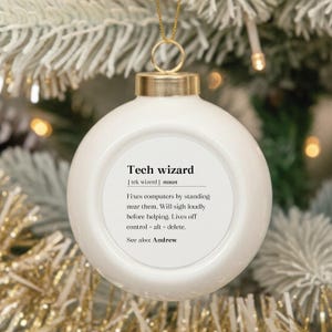 May include: A white Christmas ornament with gold accents and a gold hanging loop. The ornament has the text "Tech wizard" with a definition printed on it. The ornament is hanging on a Christmas tree.
