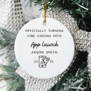 May include: A white, round Christmas ornament with gold string. The ornament has the text "OFFICIALLY TURNING VIBE CODING INTO App Launch" and "JASON SMITH" printed in black. A graphic of a rocket launching from a phone is also included.