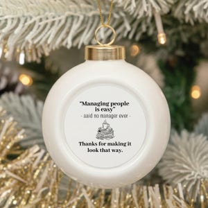 May include: A white spherical ceramic ornament with a gold-colored metal cap and hanging loop. The center features black text that reads: "Managing people is easy" - said no manager ever - followed by a small coffee cup and stack of papers icon, and "Thanks for making it look that way."