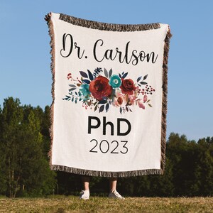 K&ouml;nnte beinhalten: Eine wei&szlig;e gewebte Wolldecke mit einem Blumendesign und dem Text "Dr. Carlson PhD 2023".
