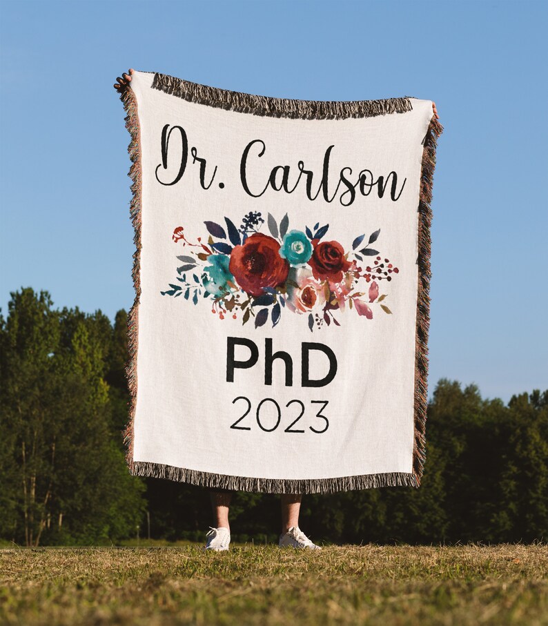 K&ouml;nnte beinhalten: Eine wei&szlig;e gewebte Wolldecke mit einem Blumendesign und dem Text "Dr. Carlson PhD 2023".