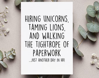 May include: A white card with black text that reads "Hiring unicorns, taming lions, and walking the tightrope of paperwork...Just another day in HR!" The card is surrounded by eucalyptus leaves and small white flowers.