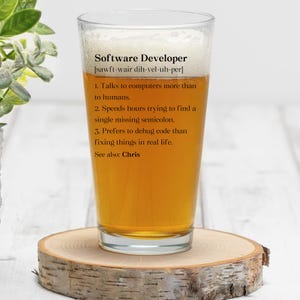 May include: A pint glass filled with beer, resting on a wooden coaster. The glass has the words "Software Developer" and a definition printed on it. The definition includes statements about computer interaction and debugging code.
