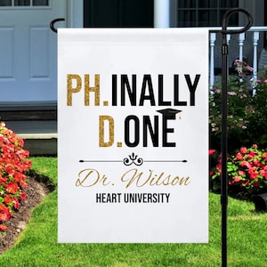 May include: White graduation yard sign with gold glitter text that reads "PH.INALLY D.ONE" with a black graduation cap and the text "Dr. Wilson HEART UNIVERSITY".