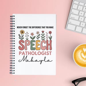 May include: A white spiral-bound notebook with the text "NEVER FORGET THE DIFFERENCE THAT YOU MAKE" and "SPEECH PATHOLOGIST Makayla" in colorful, floral-themed lettering. The notebook is on a pink surface.